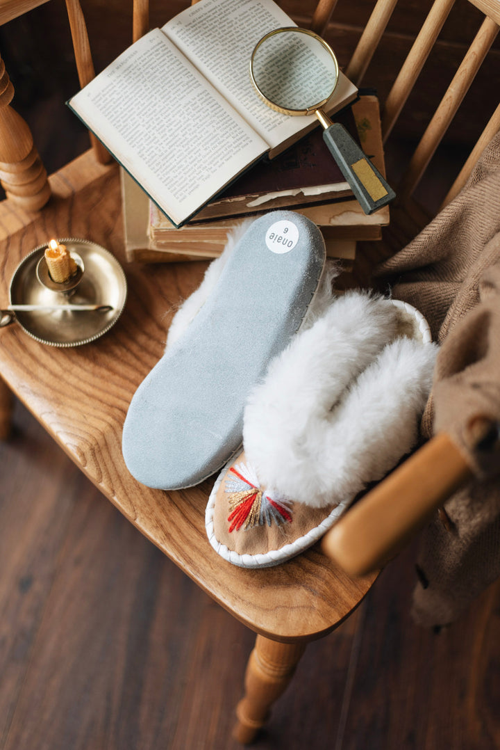 A pair of cozy slip-on slippers placed on a wooden chair, styled with an open book, magnifying glass, candle, and a brown textile in a warm indoor setting. One slipper is light blue suede-like, and the other is white faux fur with a small red tassel detail.