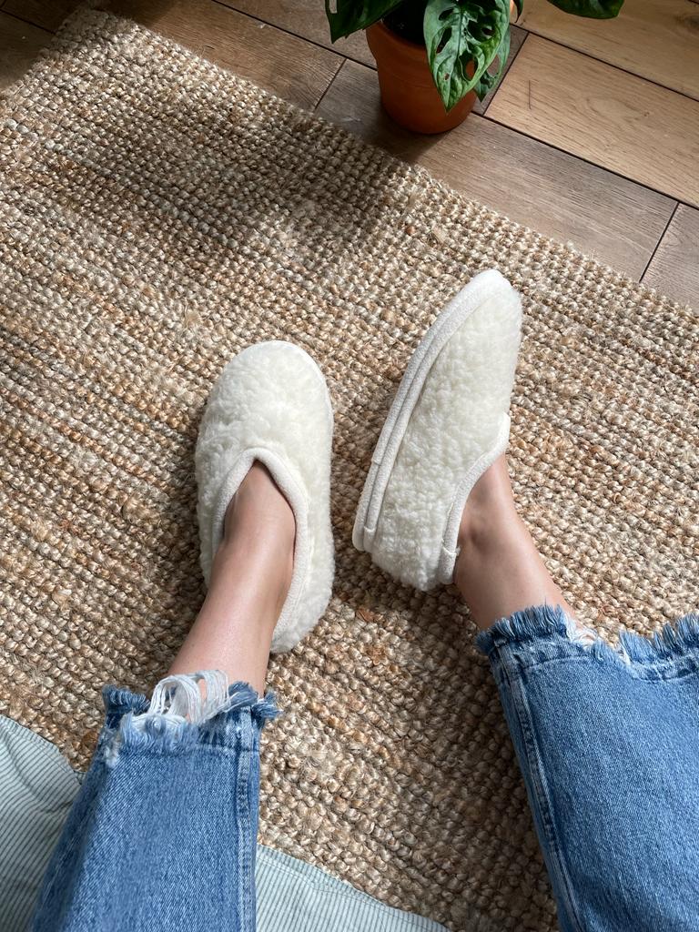 White slippers worn on a person's feet with blue jeans on a textured rug.