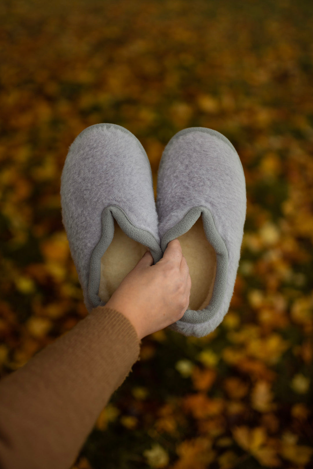 A pair of wool light gray slip-on slippers held by a hand against a blurred background of fallen autumn leaves.