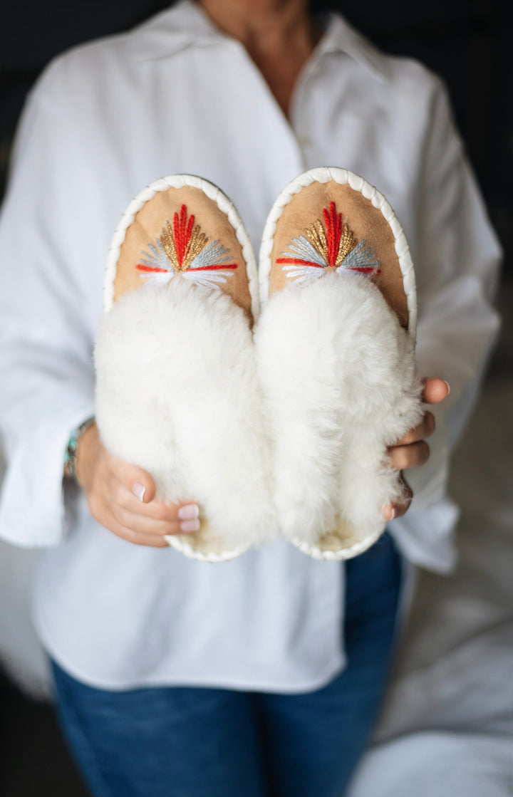 A person in a white shirt holds a pair of fluffy white slippers with colorful hand-embroidered details on the footbeds, set against a softly blurred indoor background.