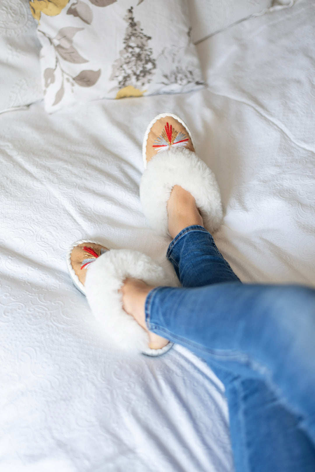 A person wearing fluffy white slippers with small red and green decorative accents, sitting on a white bed with blue jeans visible.