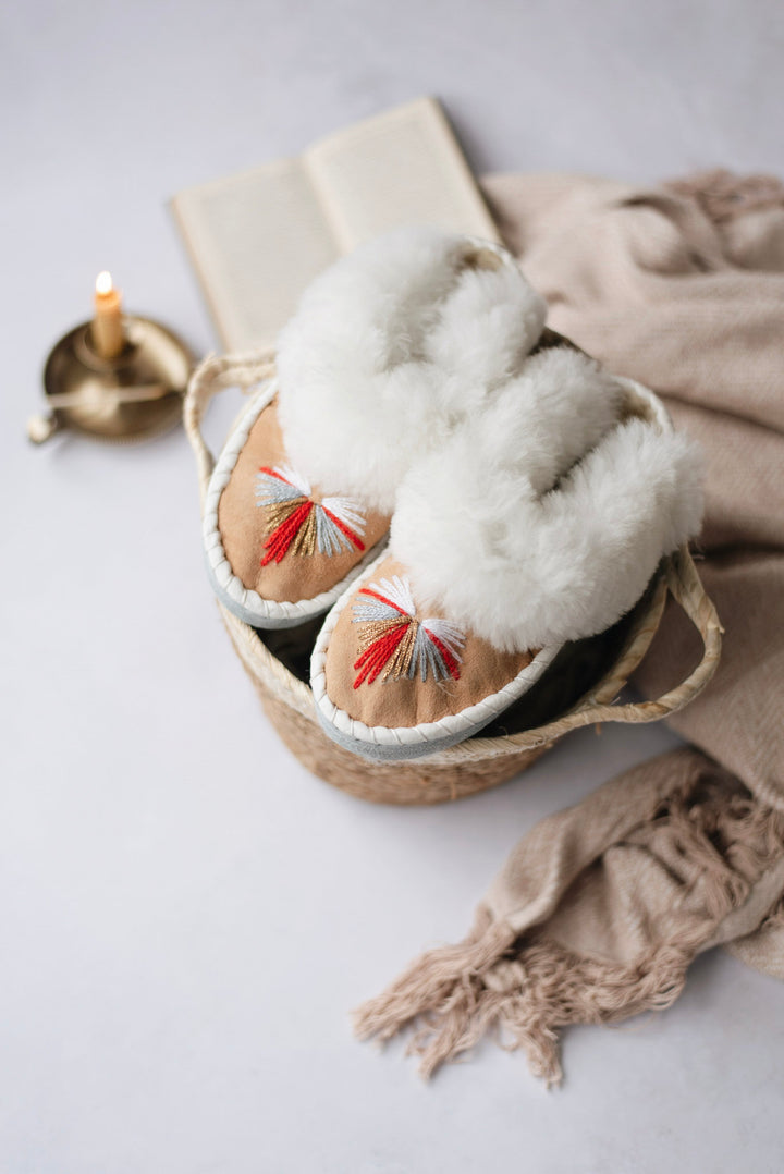 A pair of fluffy white slippers with red and multicolor embroidered details on the toes, displayed in a woven basket with a beige scarf and an open book on a light gray surface.