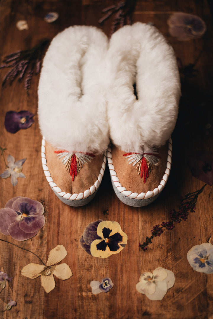 A pair of handmade moccasin-style slippers with fluffy white shearling uppers and tan suede footbeds, decorated with red and white embroidery, displayed on a rustic wooden surface with dried flowers.