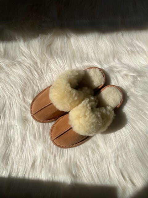 Beige wool slippers with fluffy sheepskin cuff on suede sole placed on soft white sheepskin rug