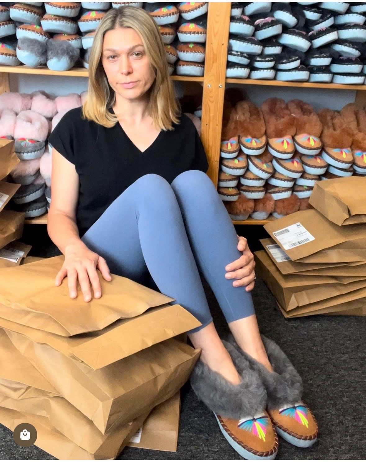 Eve sits on a stack of brown paper mailers in a storage, wearing gray sheepskin slippers with colourful embroidered details. Shelves behind her are filled with more sheepskin slippers and stacked brown mailers.