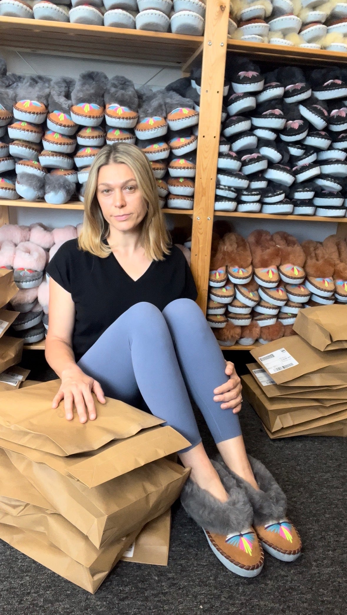 eve onaie owner sits on the floor in front of shelves filled with packaged slippers, wearing brown sheepskin slippers with colourful painted toe embroidery. She is wearing a black top and blue leggings, surrounded by brown paper mailers.