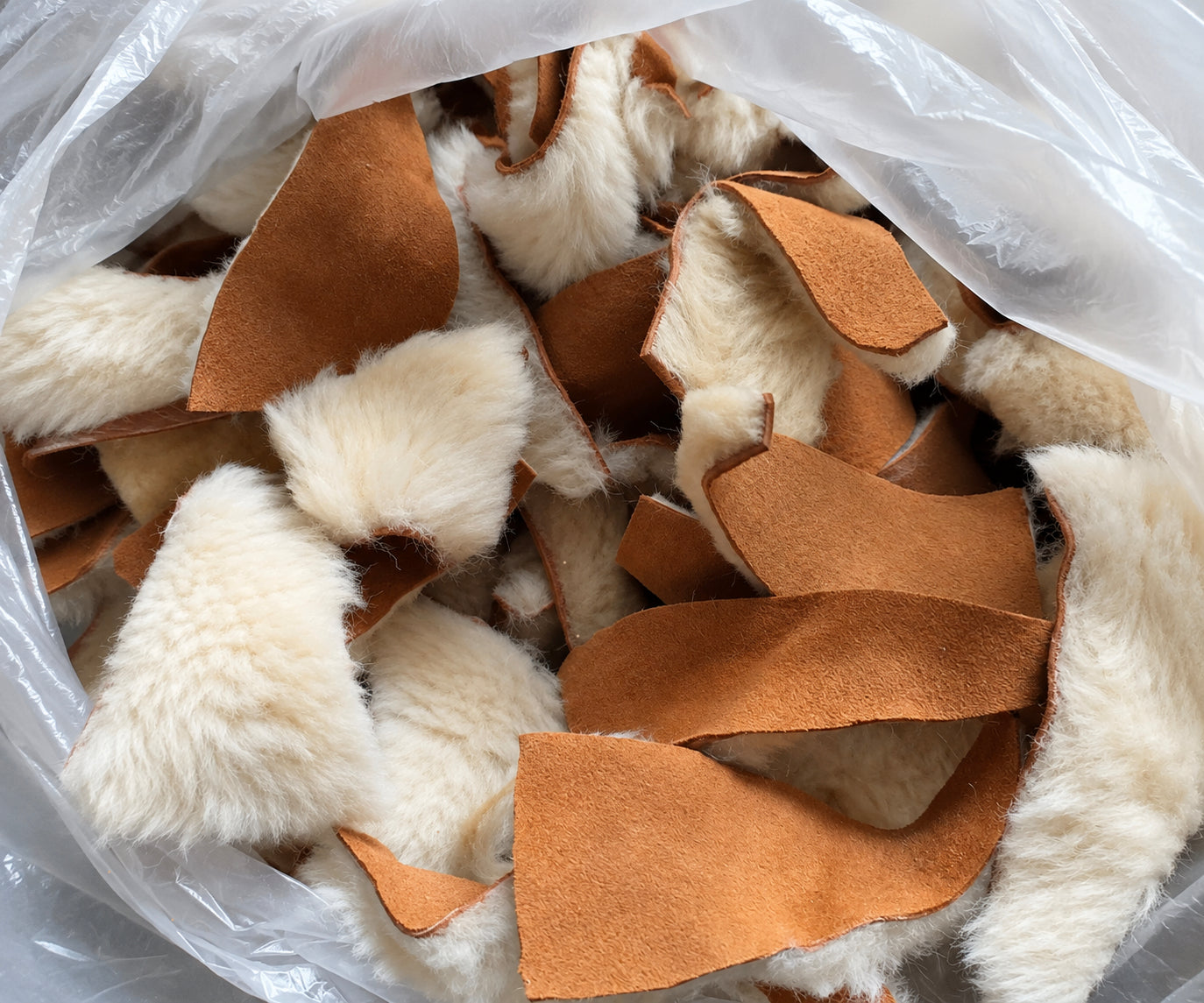 A clear plastic bag filled with cut pieces of fluffy off-white sheepskin mixed with tan suede or leather strips, photographed from above.