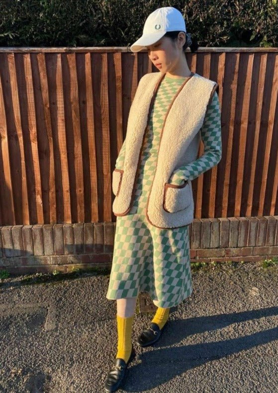 Person wearing a green patterned dress with a beige coat and white cap, standing in front of a wooden fence.