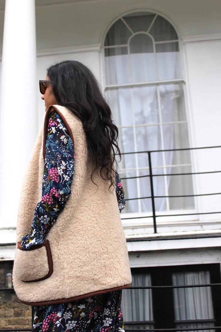 Woman wearing a beige vest over a floral dress in front of a building with large windows.