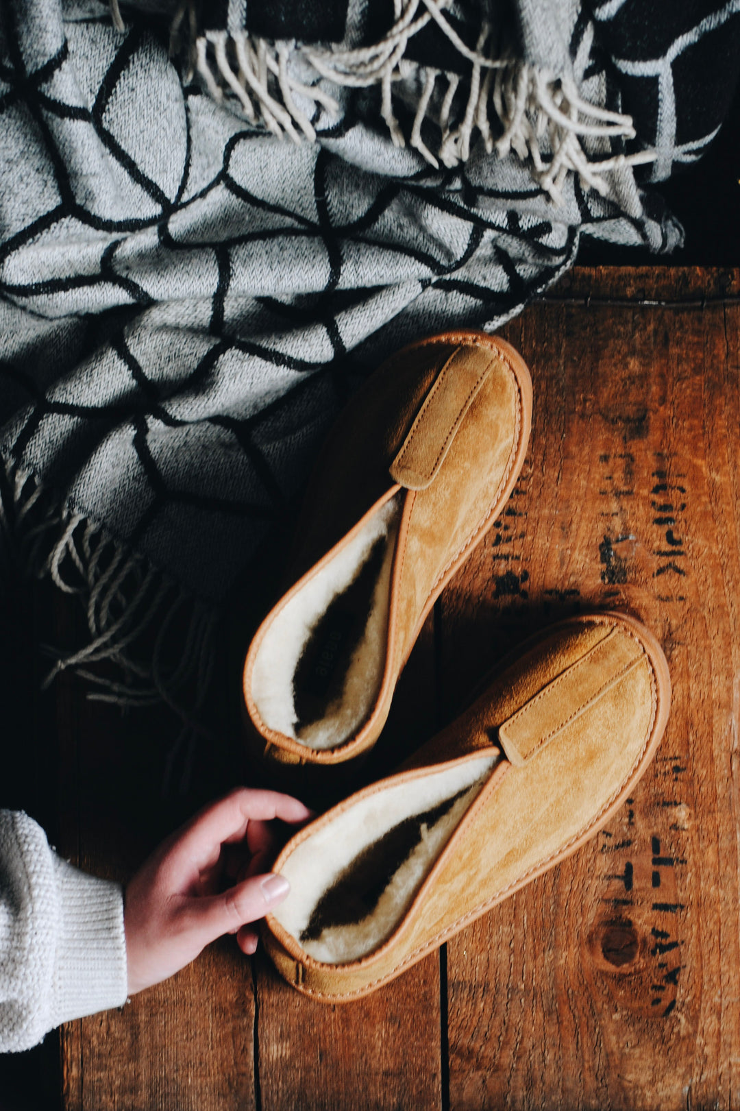 A pair of tan suede slip-on slippers with a cream shearling lining, shown on a rustic wooden table with a hand reaching in. A black and gray patterned knit blanket is draped in the background.